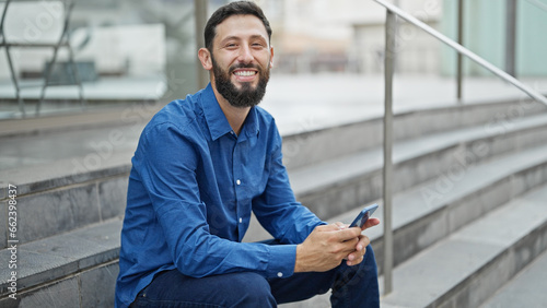 Young hispanic man business worker using smartphone sitting on stairs at street
