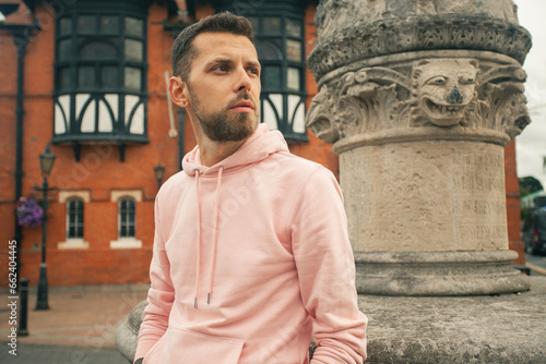 Welcome concept. Young bearded romantic man wearing pink trendy hoodie posing over brick house with flowers on backgound. Old classical fontain. Close up. Outdoor shot