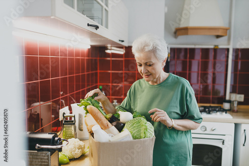 Image of senior caucasian housewife unpacking carton box with vegetables and food products after home delivery from grocery store, standing at kitchen with curious facial expression
