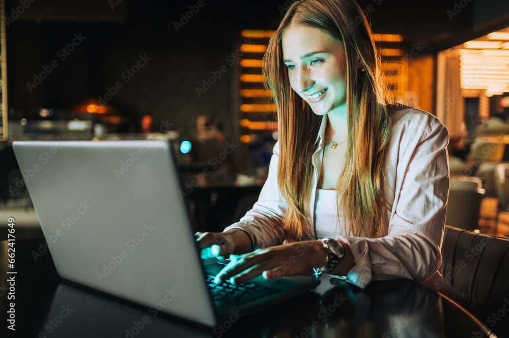 A cheerful young female entrepreneur is sitting in a cafe and typing on a laptop in the dark while smiling at the laptop. A cheerful young female entrepreneur is sitting in a cafe and typing on a laptop in the dark while smiling at the laptop.