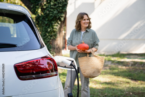 Woman with shopping bag and pumpkins next to a charging electric car in the yard of a country house