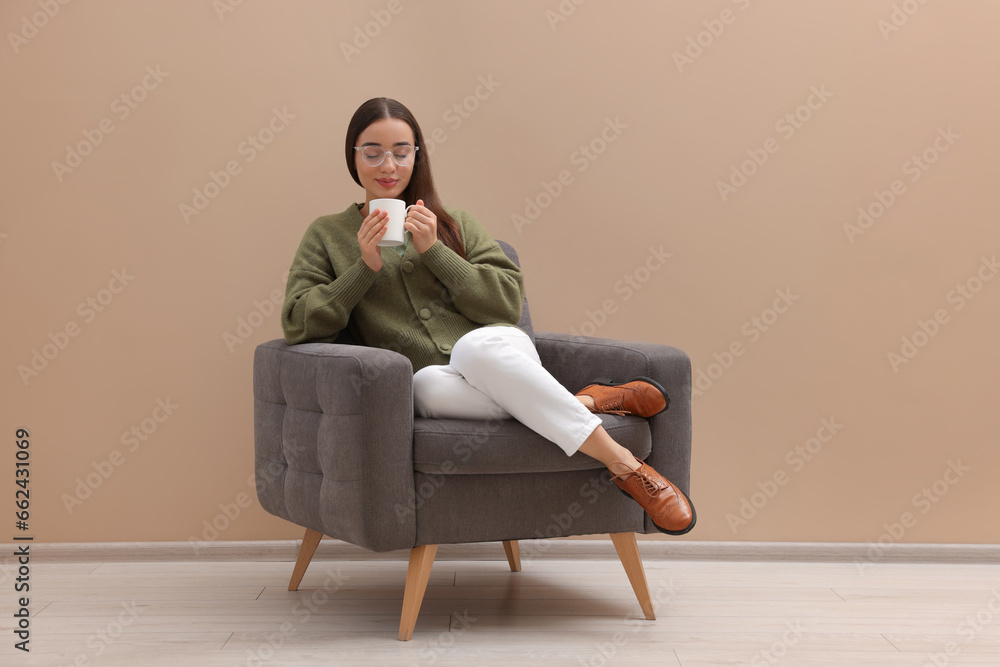 Beautiful woman with cup of drink sitting in armchair near beige wall indoors