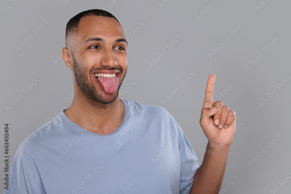 Fototapeta premium Happy young man showing his tongue and pointing at something on light grey background