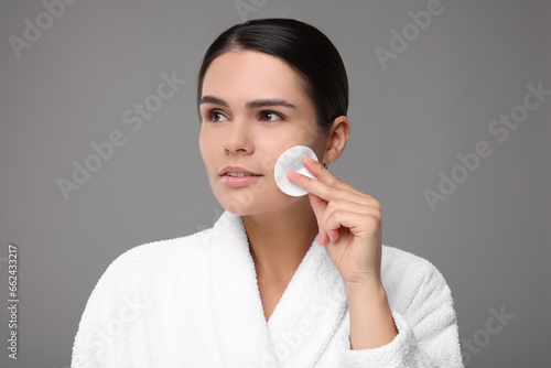 Young woman cleaning her face with cotton pad on grey background