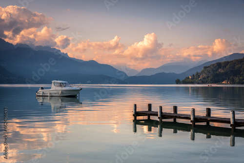 Closer view of a boat moored beside a jetty in the calm waters of Lake Annecy and the Alps mountain range in the background at sunrise, Lake Annecy, Auvergne - Rhône - Alps, France
