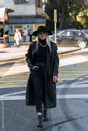 An attractive young woman crosses the road at a pedestrian crossing. A girl in stylish clothes, in a black trench coat and hat © Павел Костенко