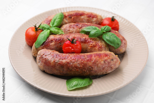 Plate with tasty homemade sausages, basil leaves and tomatoes on white tiled table, closeup