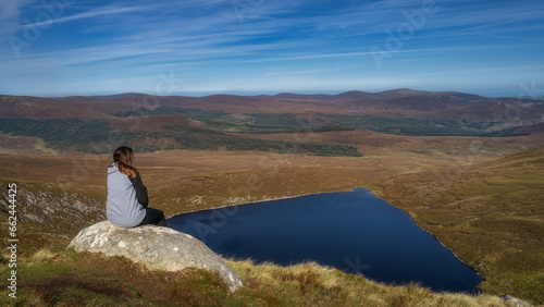 Young woman sitting on a rock and admiring a beautiful view of heart shaped lake, Lough Ouler and mountain range, Wicklow Mountains, Ireland