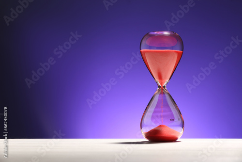 Hourglass with red flowing sand on white table against purple background. Space for text