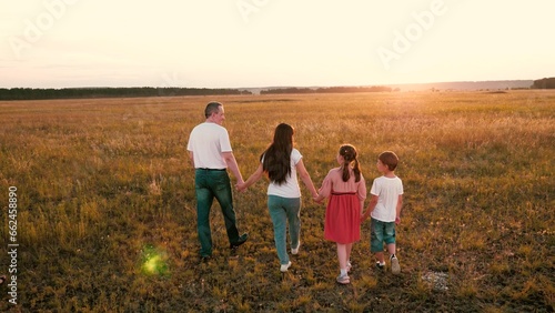 Loving parents hold hands of cute children walking across evening field