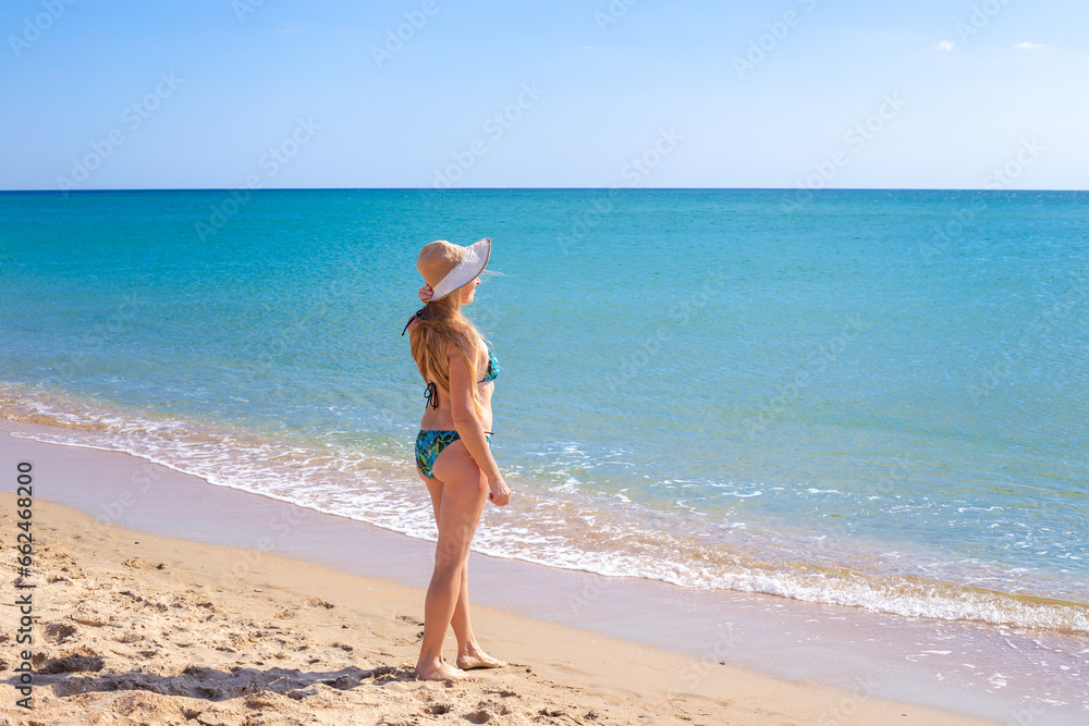 A woman with long blond hair in a swimsuit and hat stands on the shore of the blue sea and looks into the distance. Tourism and sea recreation.