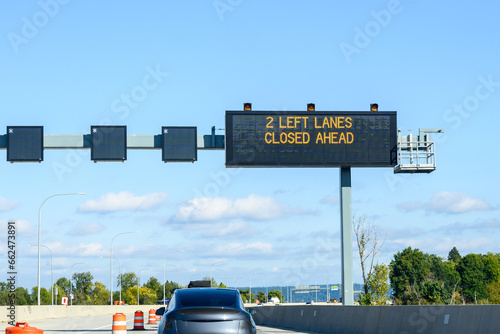 Overhead electronic highway sign, 2 left lanes closed ahead, on a sunny blue sky day
