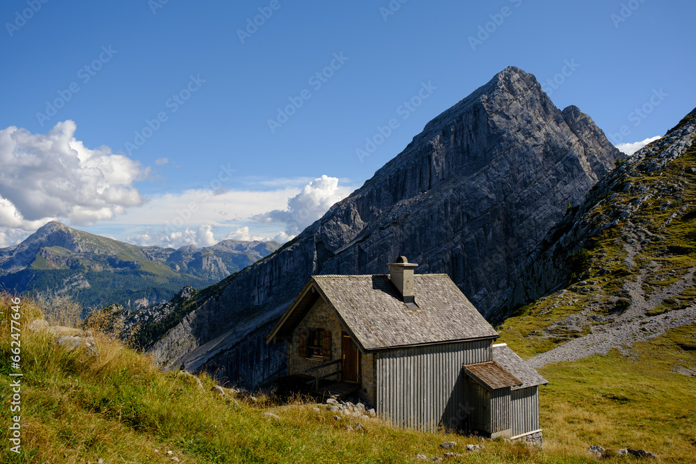 Hangkante auf Hochebene mit Bergspitze im Hintergrund und naher Hütte im Vordergrund