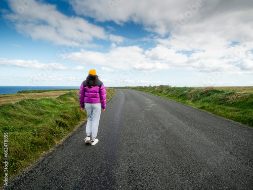 Teenager girl in colorful purple jacket and yellow hat walking on a small country road on a warm sunny day with blue cloudy sky. Travel and sightseeing theme. Outdoor activity, healthy lifestyle.