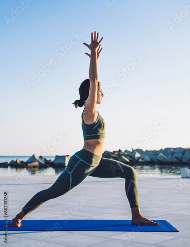 Young slim brunette making yoga Crescent lunge pose on mat against background of sky