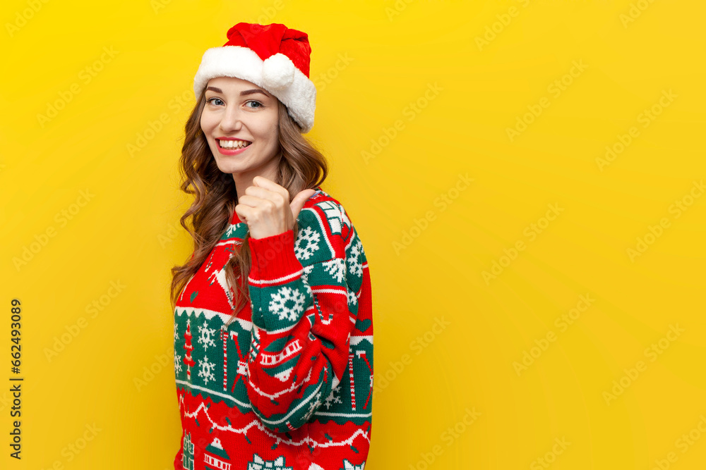 young girl in christmas sweater and santa claus hat smiles and points back on yellow isolated background young girl in christmas sweater and santa claus hat smiles and points back on yellow isolated background