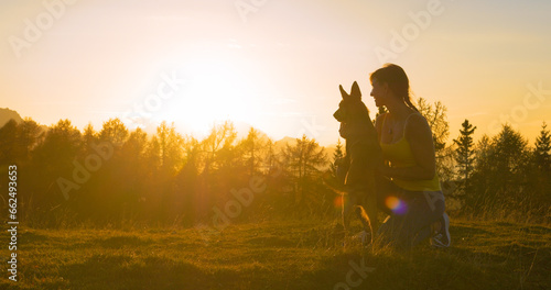 CLOSE UP, LENS FLARE: Smiling lady watches sunset from grassy peak with her dog