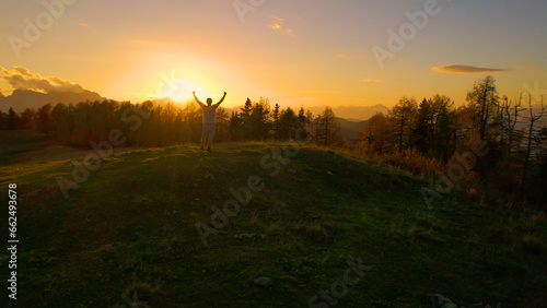 AERIAL: Happy man with raised arms standing on top of a hill in golden light