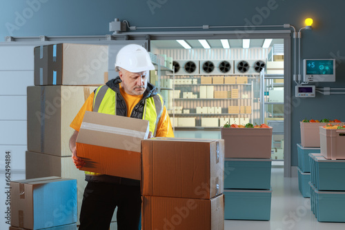 Man warehouse worker. Industrial refrigerator behind man. Man loader takes boxes out refrigerator chamber. Warehouse worker near refrigeration chamber. Refrigerator for storing fruits and vegetables
