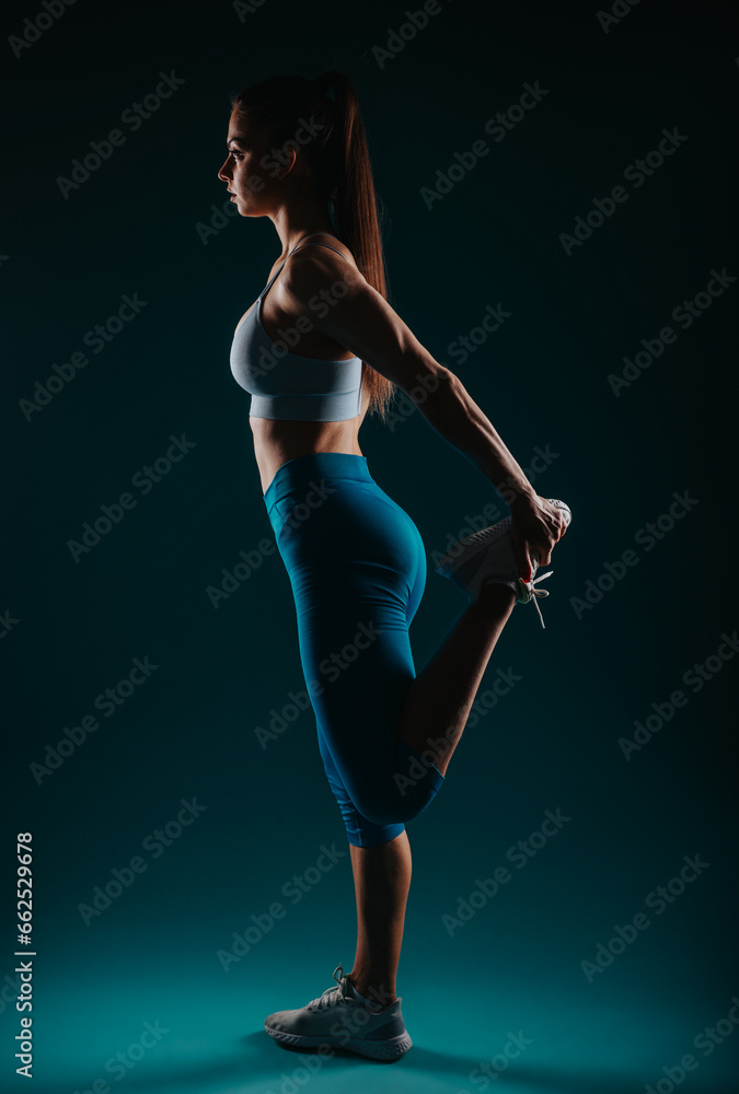 Confident female fitness model posing in a dark studio, showcasing her strong muscles and transformation.