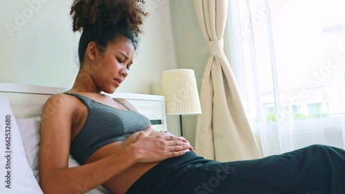 Pregnant african american mother sitting on the bed looking at her baby gently stroking her womb as her baby moves violently causing labor pains : Mothers takecare their pregnancy health at home. photo