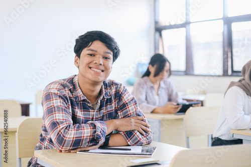Smiling young man at college leaning on desk with classmates in background.
