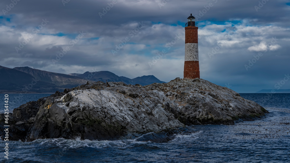 The southernmost old lighthouse Les Eclaireurs on the edge of the world. A tall red-and-white striped tower stands on a small rocky island in the Beagle Channel. The waves are foaming on the cliff.