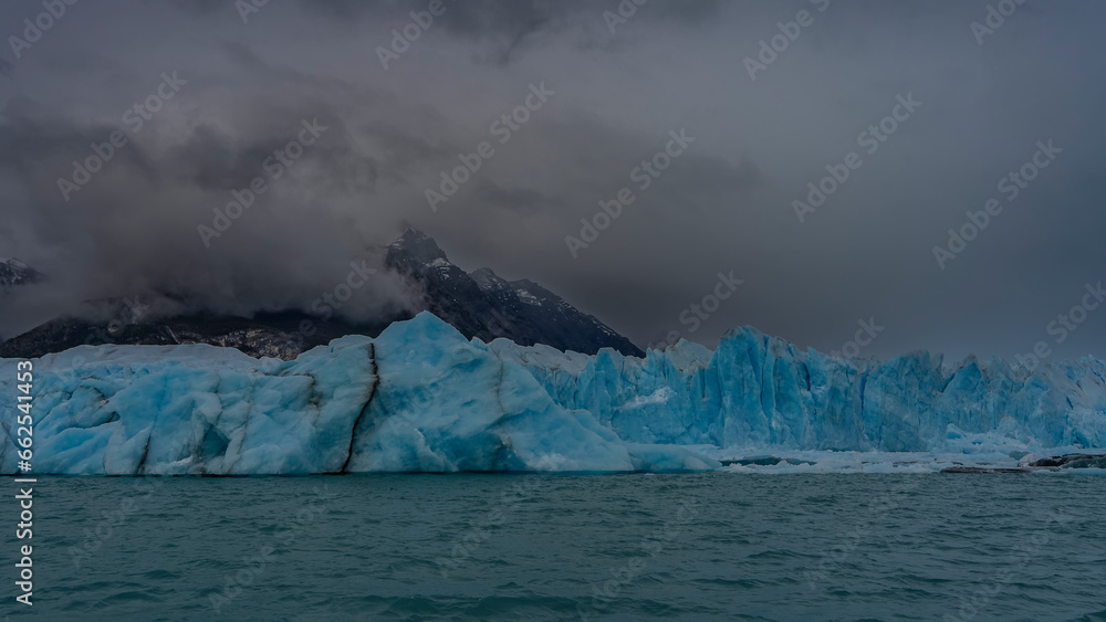 The beautiful blue glacier Perito Moreno rises above the turquoise lake. A wall of ice with cracks, crevices, sharp peaks against the mountains and a cloudy sky. El Calafate. Argentina.