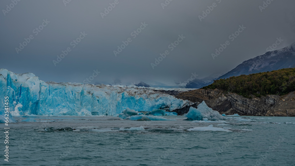 An impressive wall of blue ice with cracks and crevices. Icebergs and melted ice floes float in a turquoise lake. The layered structure of coastal rocks overgrown with forest is visible. Cloudy