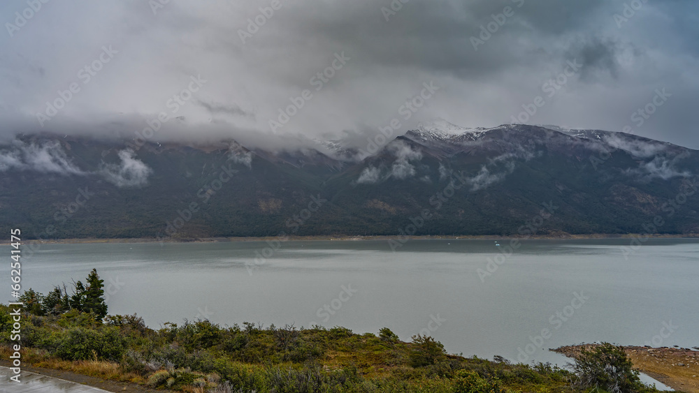 Floating blue ice floes and icebergs are visible in the glacial lake. Coastal snow-capped mountains hide in the fog. Cloudy. Lago Argentino. El Calafate. Los Glaciares National Park