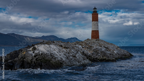The southernmost old lighthouse Les Eclaireurs on the edge of the world. A tall red-and-white striped tower stands on a small rocky island in the Beagle Channel. The waves are foaming on the cliff.