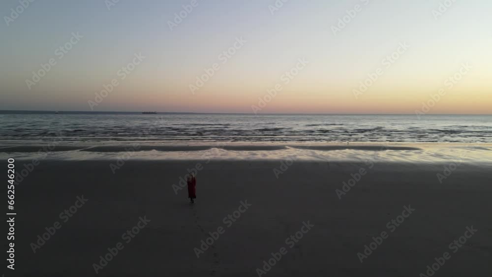Blonde woman walking on the beach with a long sweater on board as the sun rises and the drone surrounds her in a fabulous landscape. Blonde woman walking on the beach with a long sweater on board as the sun rises and the drone surrounds her in a fabulous landscape.