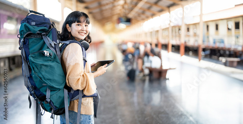 Young female traveler with backpack and using tablet, standing at train station.