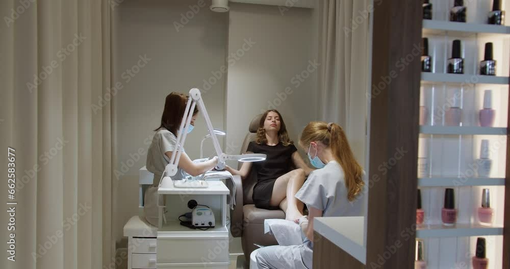 Two masters doing manicure and pedicure for female client at luxury salon. Woman relax on beauty treatment procedure for nail care at studio with great service. Selfcare concept Two masters doing manicure and pedicure for female client at luxury salon. Woman relax on beauty treatment procedure for nail care at studio with great service. Selfcare concept