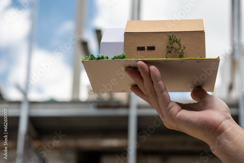 Man's hand holding a model of a dream house to compare with the house being built