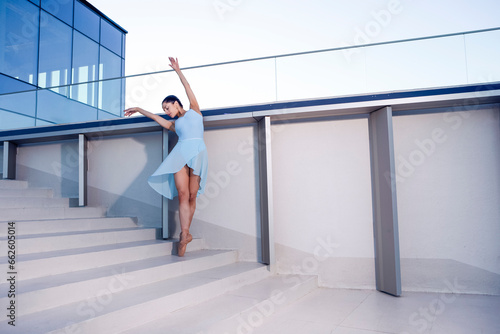 Graceful Slim Professional Caucasian Ballet Dancer in Blue Tutu Dreass Whie Posing Against White Wall In Leg Muscle Stretching Pose On Stairs In Stretching photo