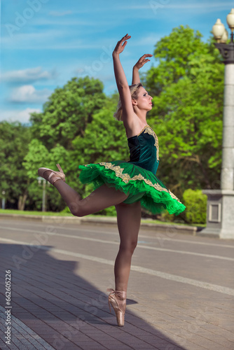 One Winsome Professional Caucasian Ballet Dancer in Green Tutu Dress Posing in Standing Position With Lifted Hands And Stretched Leg Against Blue Sky