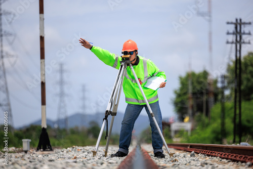 Engineer asian male wearing green uniform and orange hard hat use theodolite equipment and holdint data paper surveying construction worker on Railway site.