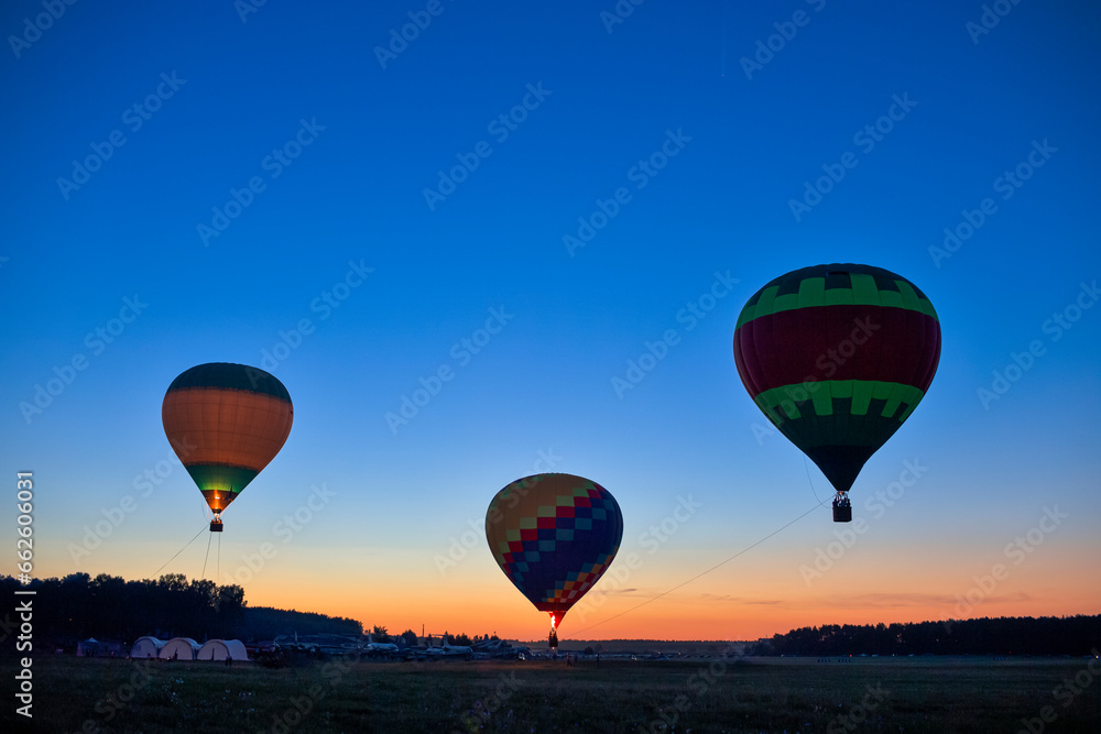 Obraz premium Line of Three Colorful Air Balloons Levitating Over the Field Outdoors Against Clear Blue Skies At Twilight.