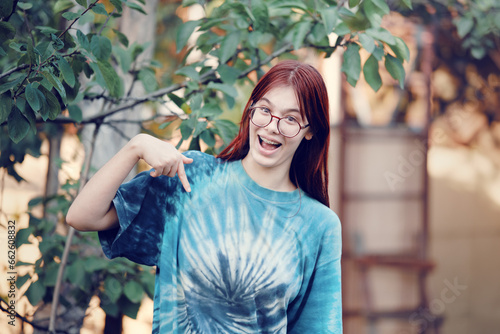 Happy red-haired teenage girl with glasses in a blue T-shirt points with her finger.