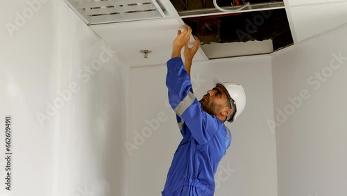 Handsome caucasian electrician man wearing safety workwear checking and repairing Maintenance light bulbs, wiring and fire sprinkler in the ceiling. Install the lighting system in the ceiling in room.