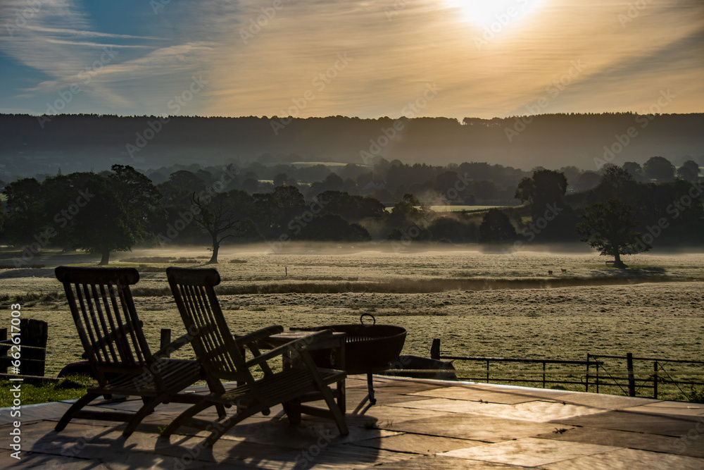 Reclining chairs, sit, and relax at a beautiful countryside view of a glorious sunrise over grassy rural landscape in Devon UK. Cattle grazing in the distance with a mist hanging over the fields. 