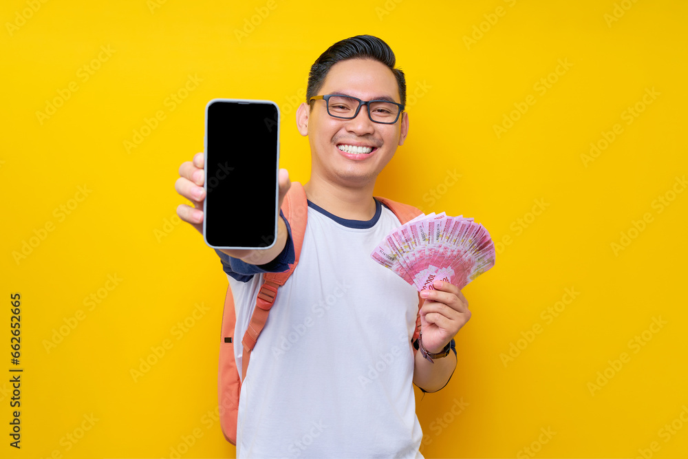 Smiling young Asian student man in casual clothes and glasses backpack holding smartphone blank screen and cash money isolated on yellow background. Education in High School University College concept Smiling young Asian student man in casual clothes and glasses backpack holding smartphone blank screen and cash money isolated on yellow background. Education in High School University College concept