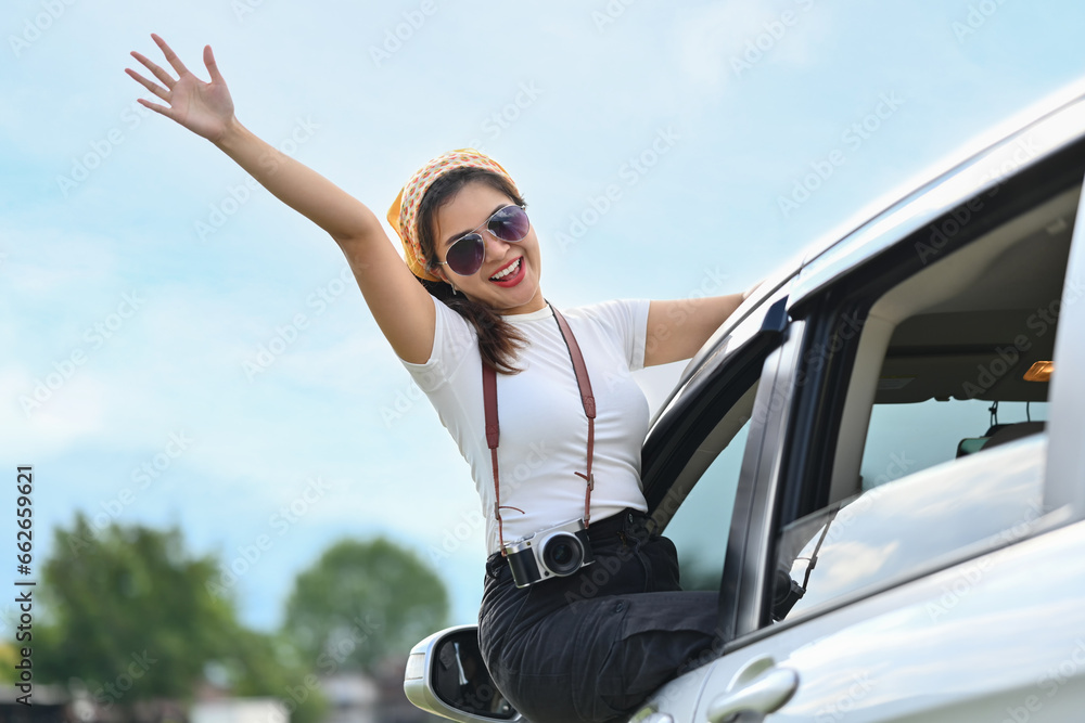 Overjoyed young woman in sunglasses leaning out from car window and waving hand during summer road trip Overjoyed young woman in sunglasses leaning out from car window and waving hand during summer road trip