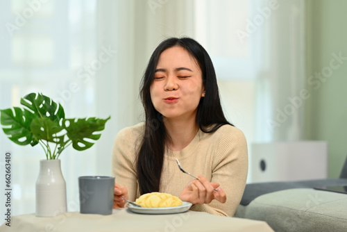 Happy young asian woman sitting in living room and eating tasty pie with closed eyes. People, food and lifestyle © Prathankarnpap