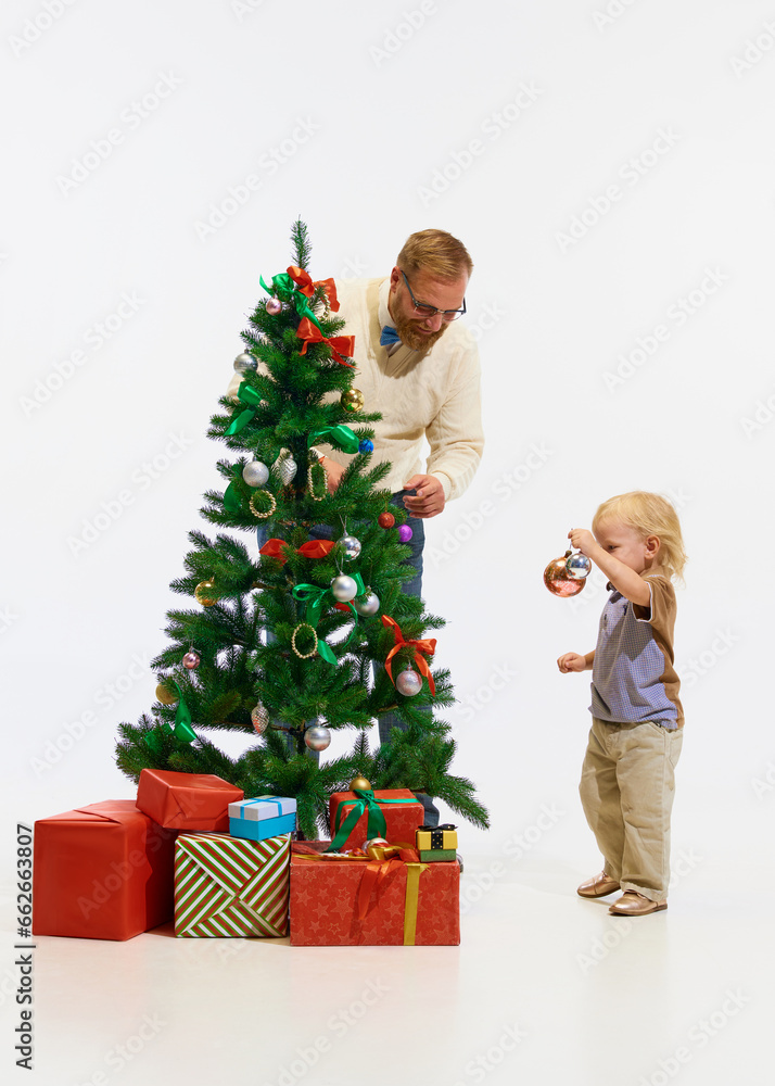 Happy winter holidays. Happy little kid, boy with father, man decorating with toys Christmas tree against white studio background. Happy winter holidays. Happy little kid, boy with father, man decorating with toys Christmas tree against white studio background.