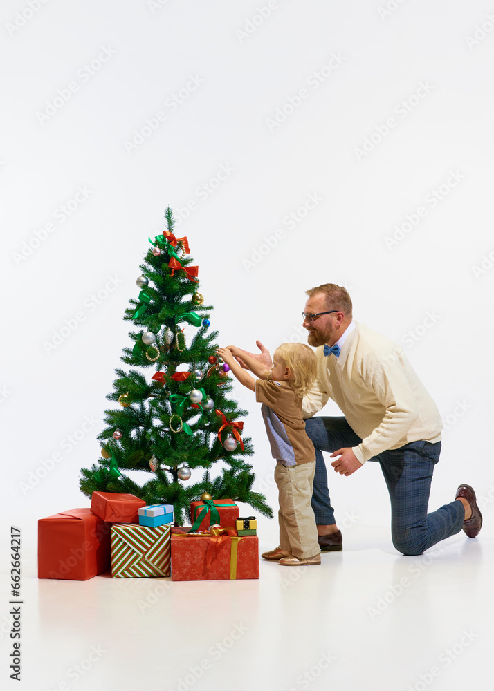 Happy winter holidays. Cheerful little kid, boy with father, man decorating with toys Christmas tree against white studio background. Happy winter holidays. Cheerful little kid, boy with father, man decorating with toys Christmas tree against white studio background.