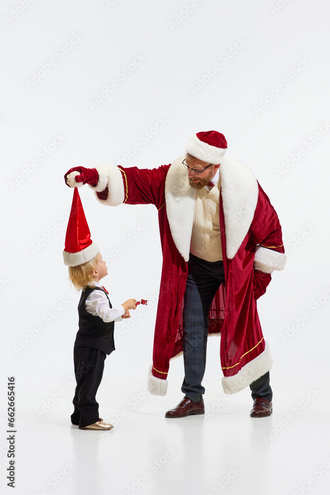 Merry Christmas. Funny father, man, dressed like Santa Claus have fun with little sweaty kid, sun against white studio background.