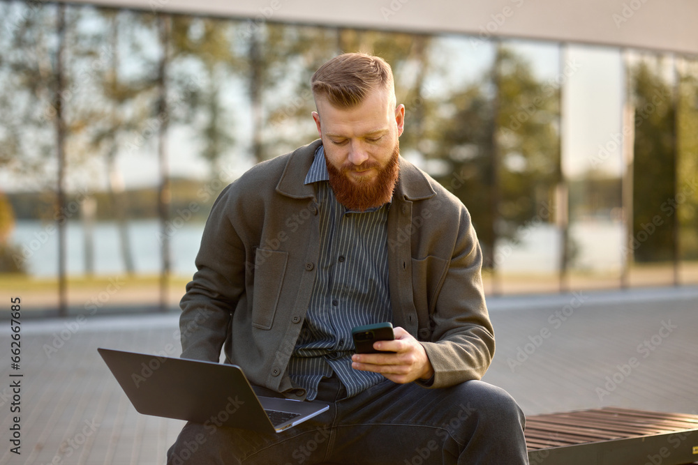 Fototapeta premium A 35-year-old man with a red beard sits on a bench with a laptop during lunch break