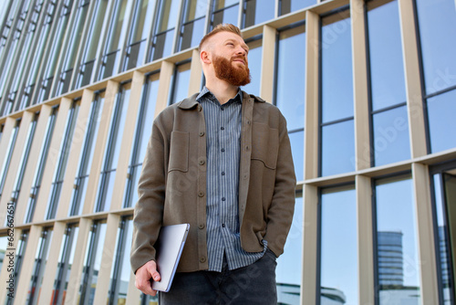 A handsome red-haired freelancer man with a beard stands confidently against the backdrop of a city building and looks into the distance